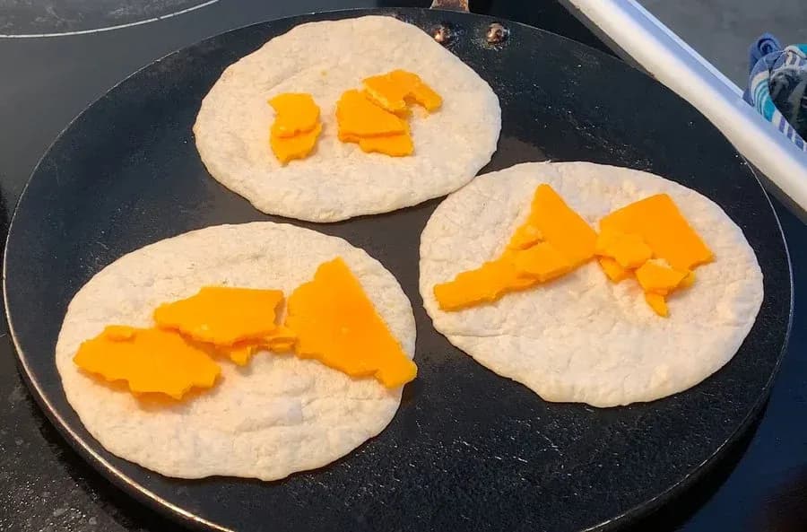 Three flour tortillas on a griddle with melted cheddar cheese strips arranged on each one.