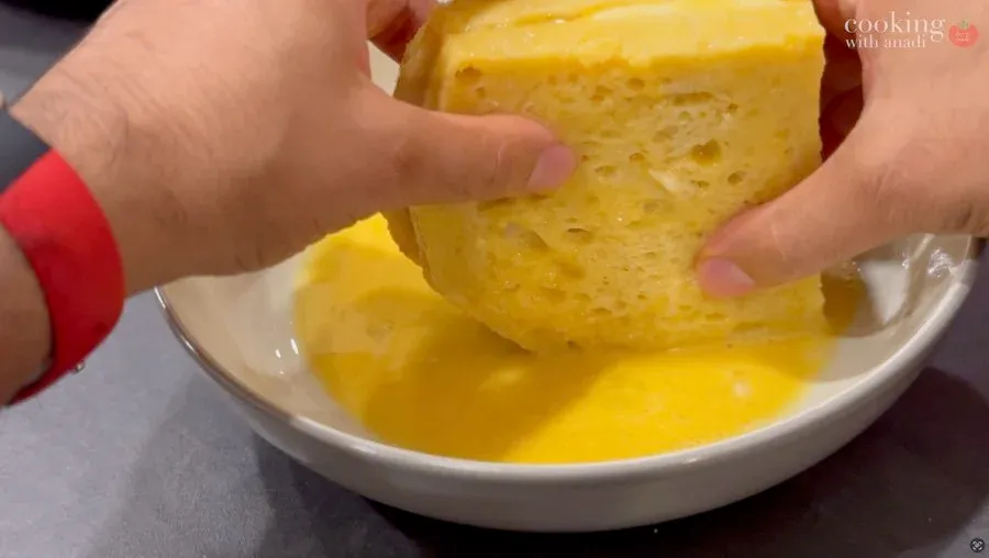 Hand dipping a breaded mozzarella in carrozza sandwich into beaten egg in a white bowl.