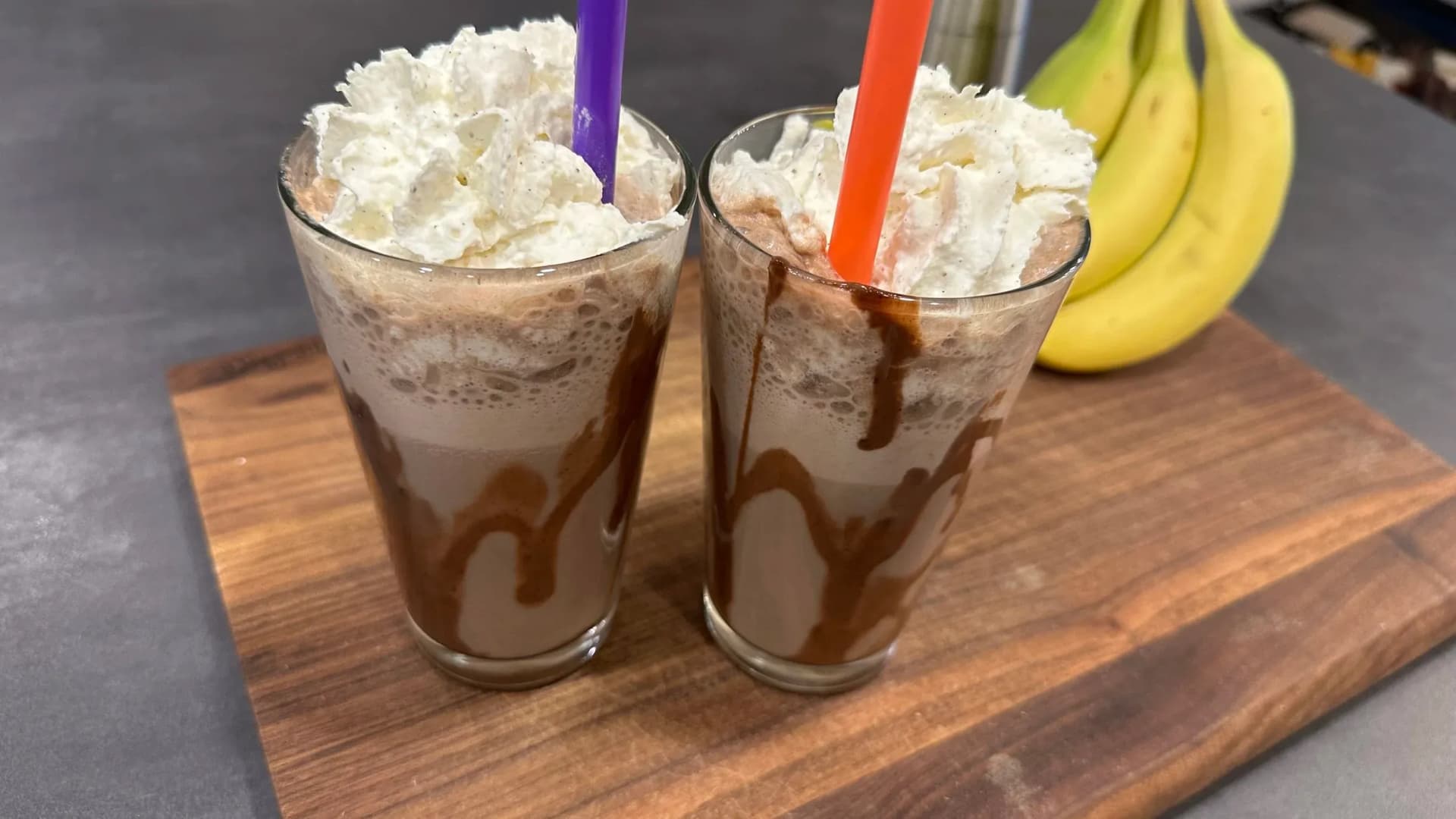 Two tall glasses of banana mocha frappe topped with whipped cream and chocolate drizzle, served with straws and fresh bananas nearby.