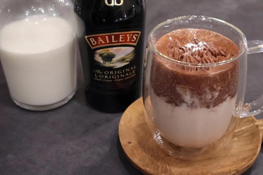 Baileys hot chocolate with whipped cream topping, served in a clear mug next to milk and Baileys bottle.