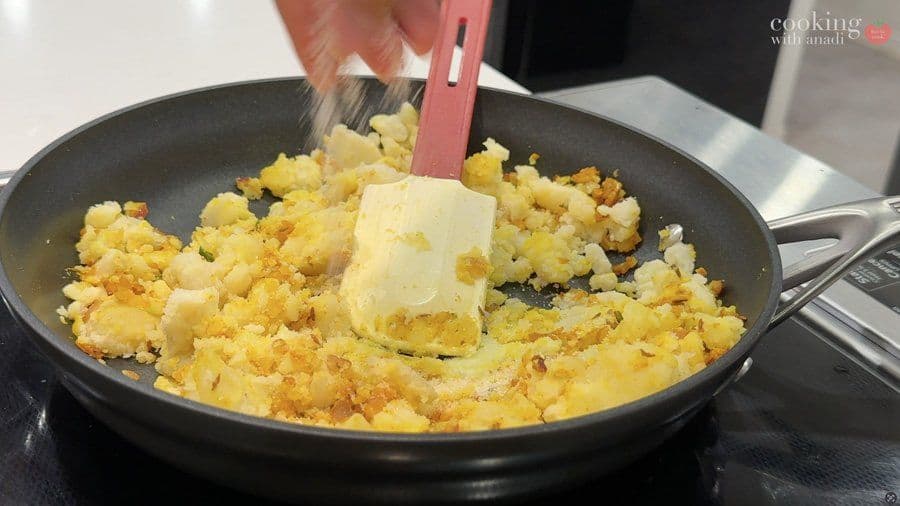 Butter being added to cooked diced potatoes and spices in a dark pan with a spatula.