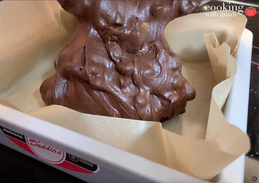 Chocolate brownie batter being spooned into a parchment-lined baking pan.
