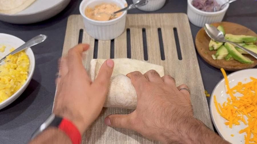 Hands rolling a flour tortilla filled with breakfast ingredients on a wooden cutting board surrounded by bowls of corn, shredded cheese, and other fillings.