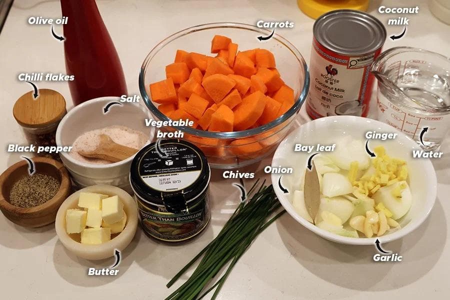 Ingredients for carrot and ginger soup laid out on a white surface including carrots, ginger, garlic, onion, butter, broth, coconut milk, and spices.