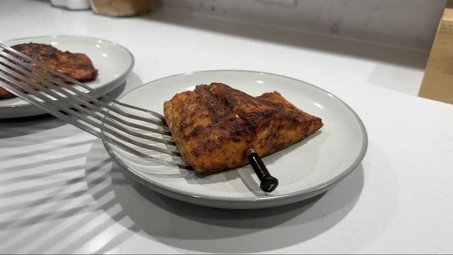 Cooked air fryer salmon fillet on a white plate with fork, showing golden-brown seasoned skin.