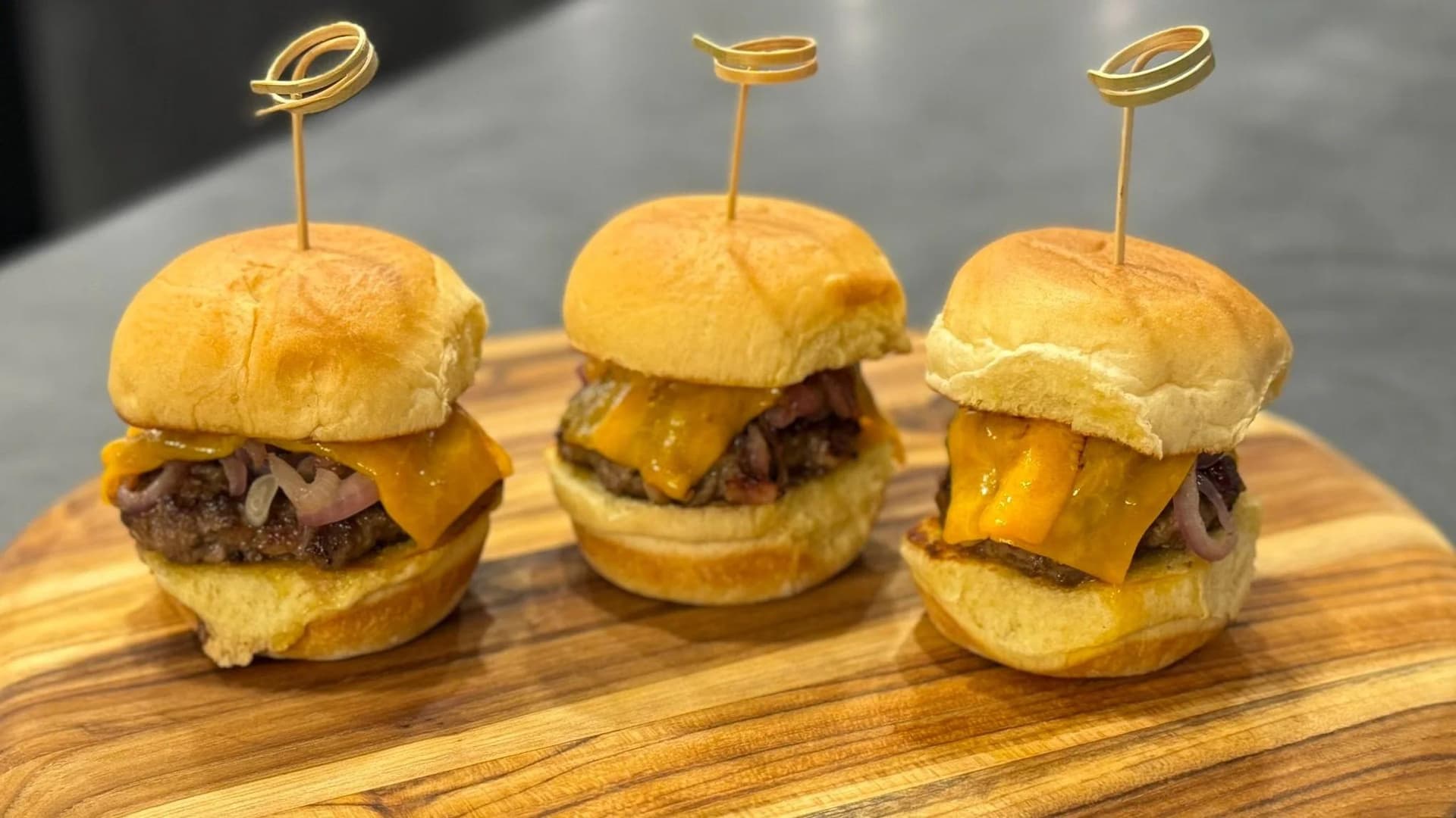 Three mini cheeseburger sliders with melted cheese and beef patties on golden buns, garnished with decorative picks, arranged on a wooden board.