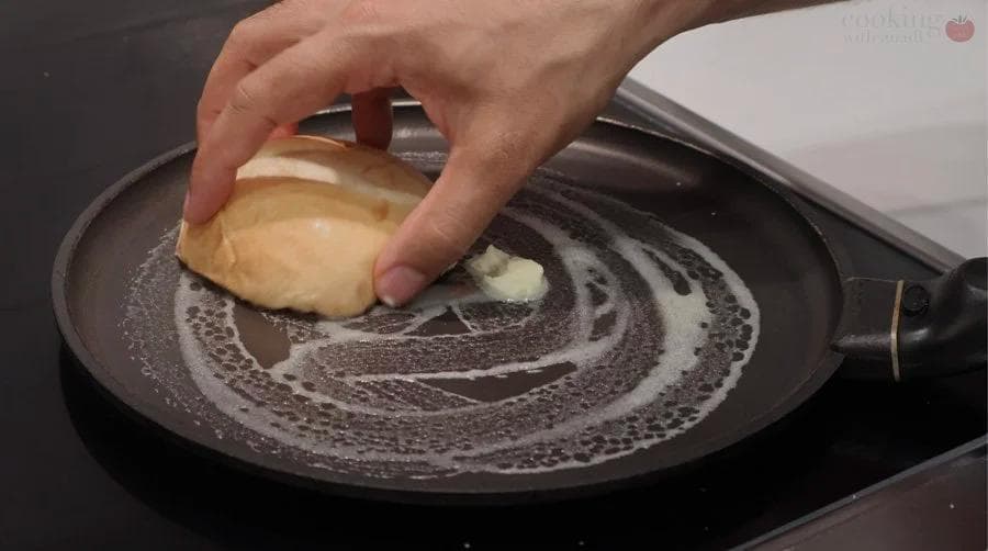 Hand spreading crab salad mixture onto a toasted bun in a skillet with melted butter.