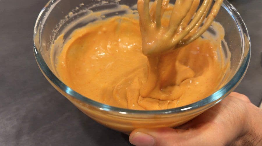 Orange spicy umami sauce mixed in a glass bowl with a whisk lifting a portion of the creamy mixture.