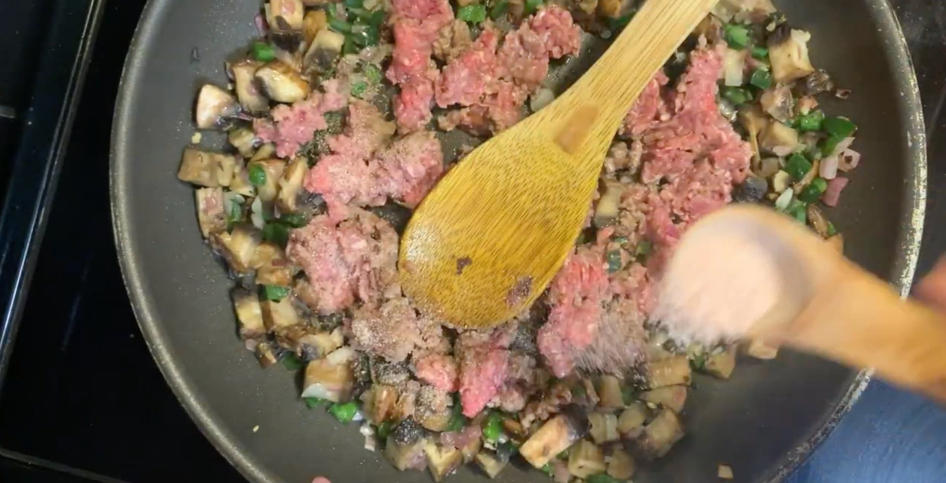 Ground beef browning in a pan with diced mushrooms and fresh herbs, being stirred with a wooden spoon.
