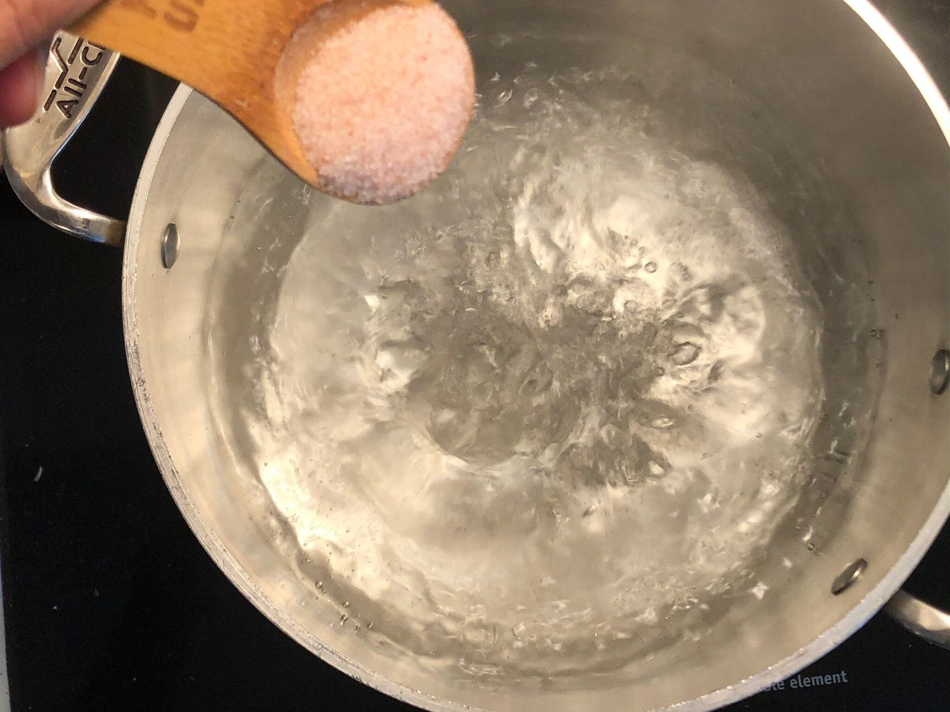 Salted boiling water in a pot with a wooden spoon adding pink salt for pasta cooking.