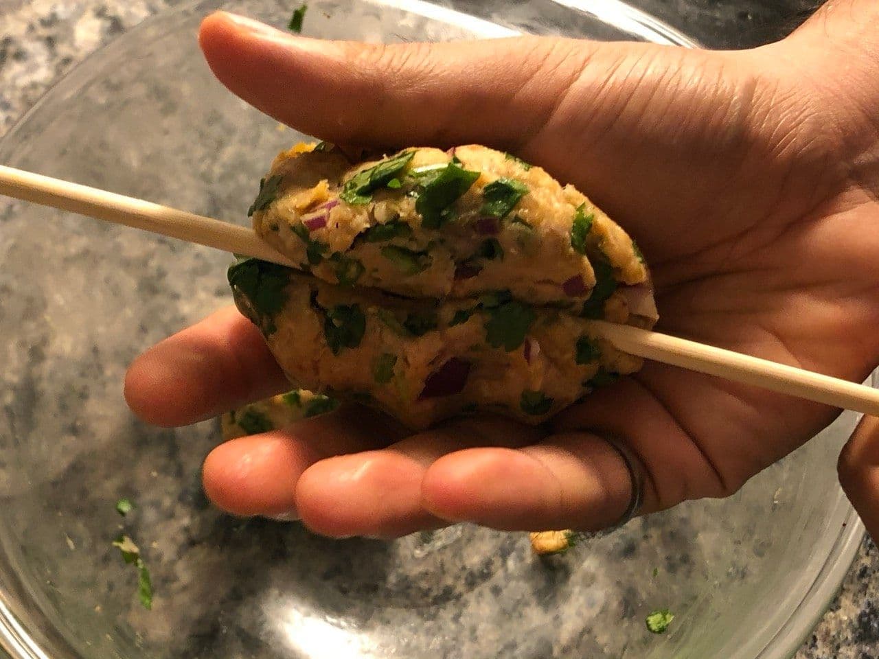 Hand holding a formed chicken seekh kebab on a skewer, garnished with green herbs and red onion pieces.