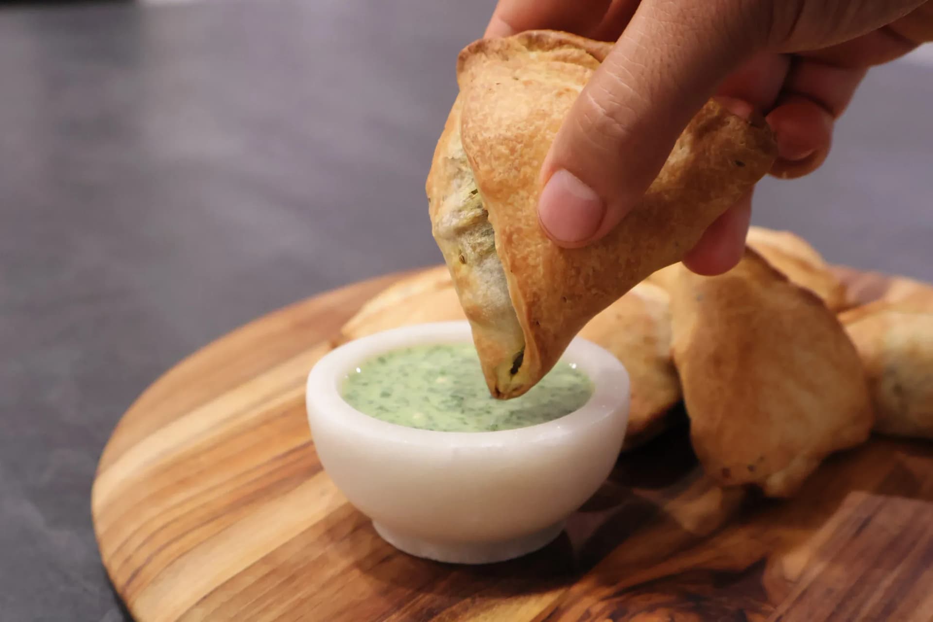 Hand dipping a golden air fryer samosa into green cilantro chutney in a white bowl on a wooden board.