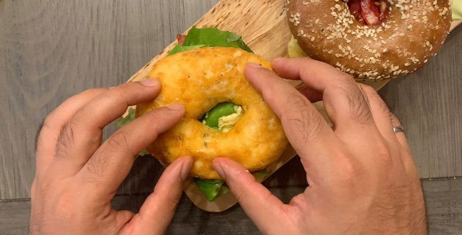 Hands assembling an avocado bagel sandwich, spreading mashed avocado and adding green pepper slices to a toasted bagel half.