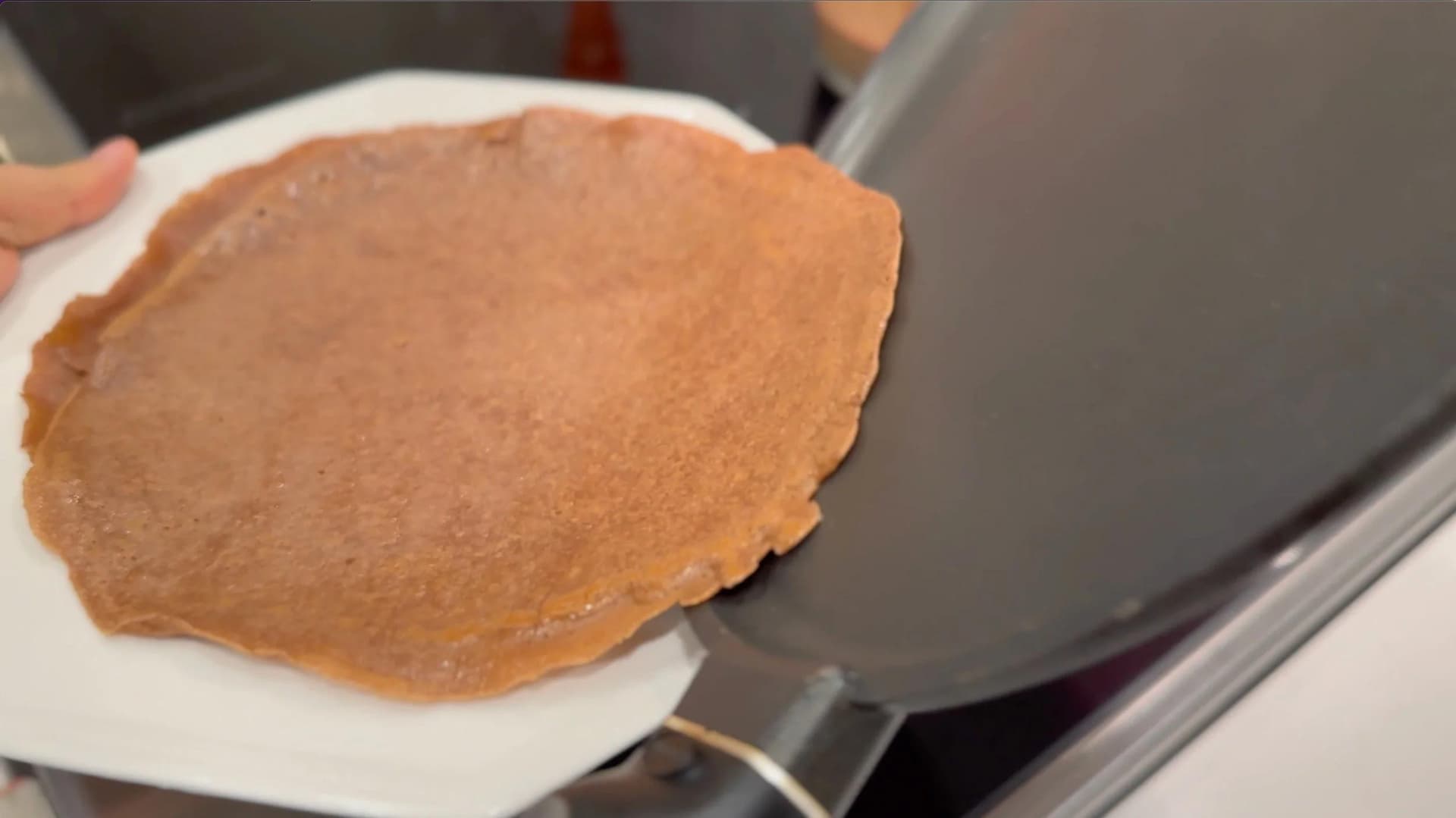 Cooked gluten-free chocolate crepe on a white plate being held next to a non-stick skillet.