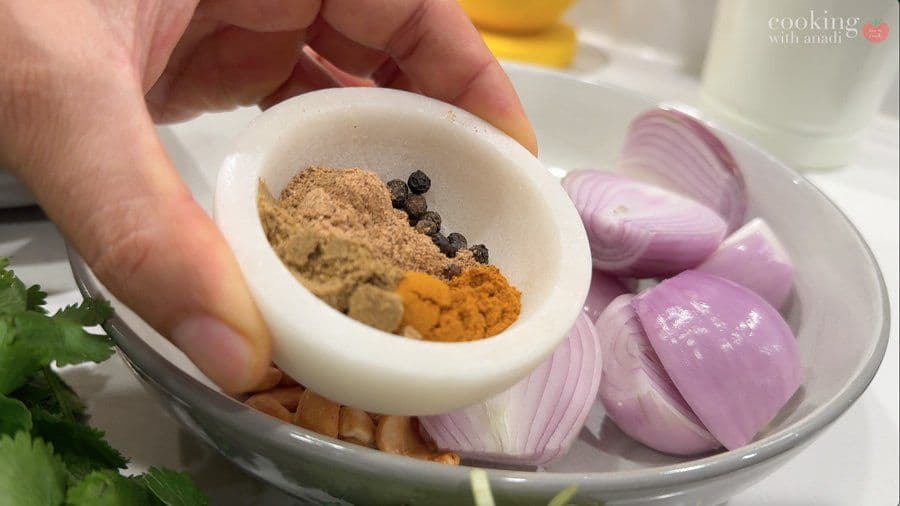 Hand holding a mortar bowl with ground spices including turmeric and black peppercorns, with sliced red onions and cilantro on plate below.