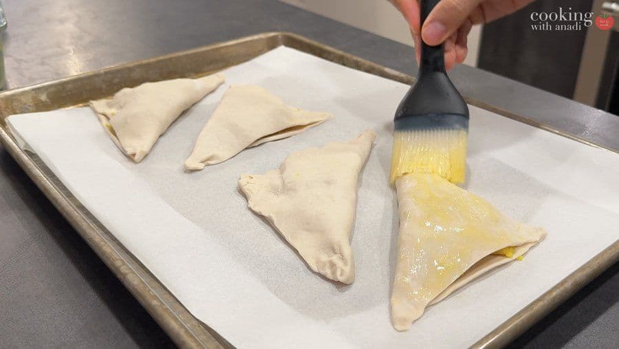 Brushing egg wash onto folded potato puff pastry triangles on parchment paper before air frying.