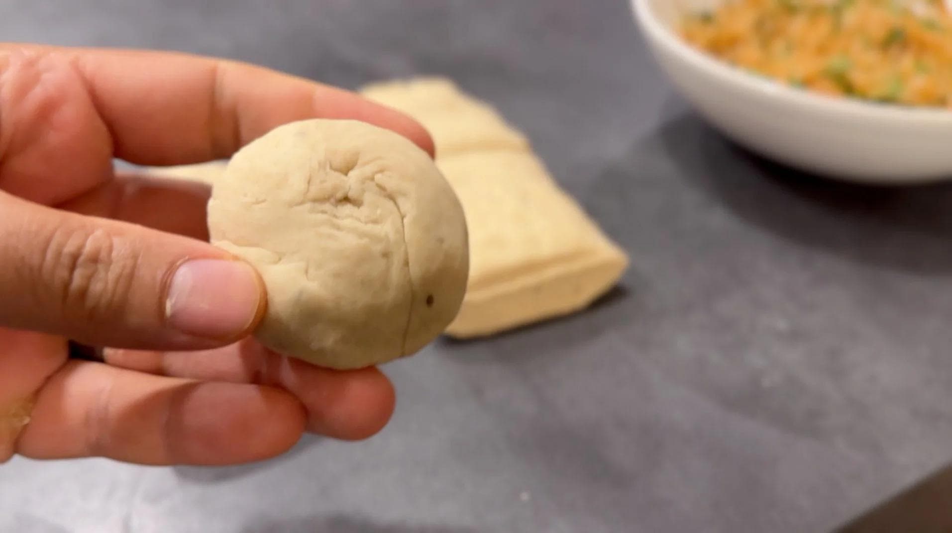 Hand holding a smooth ball of dough for gobi paratha, with dough portions and filling bowl in background.