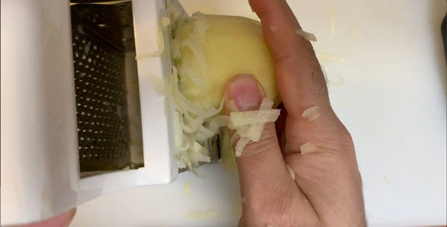 Hand holding grated potato being squeezed dry over a bowl with a grater nearby.