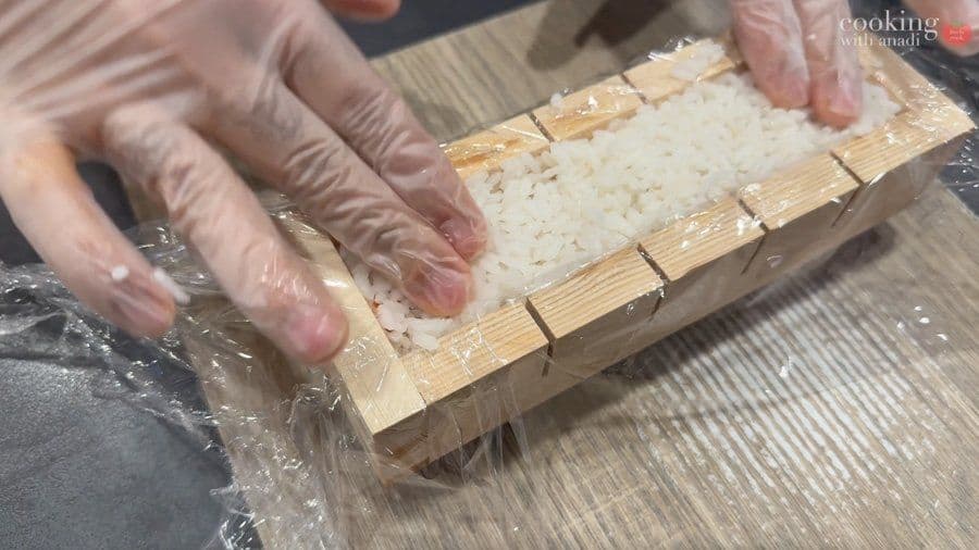 Hands spreading cooked rice into a wooden sushi mold lined with plastic wrap, preparing oshizushi.