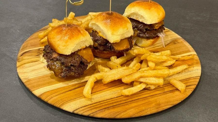 Three smash burger sliders on golden dinner rolls with melted cheese, served with crispy fries on a wooden board.