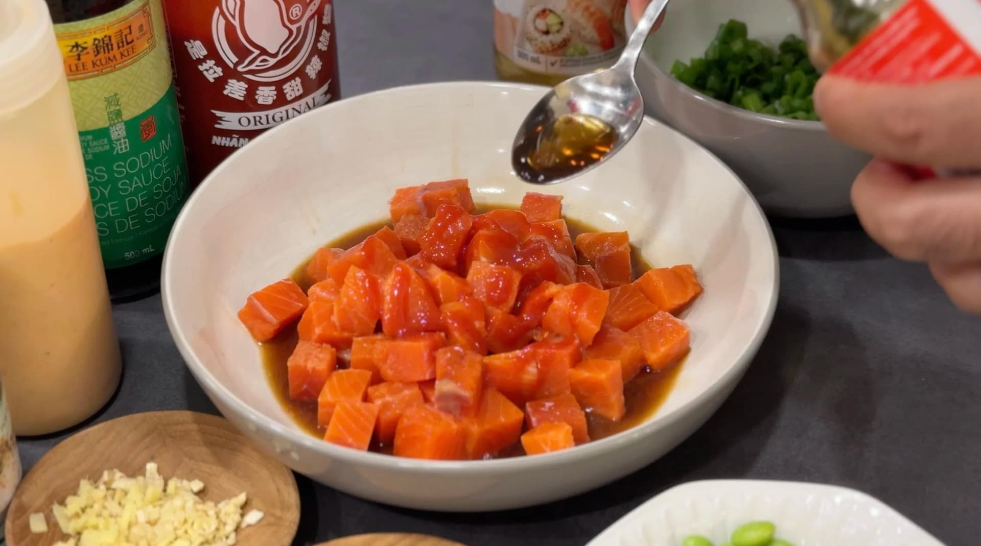 Diced salmon being drizzled with sauce in a white bowl with soy sauce and sriracha bottles nearby.