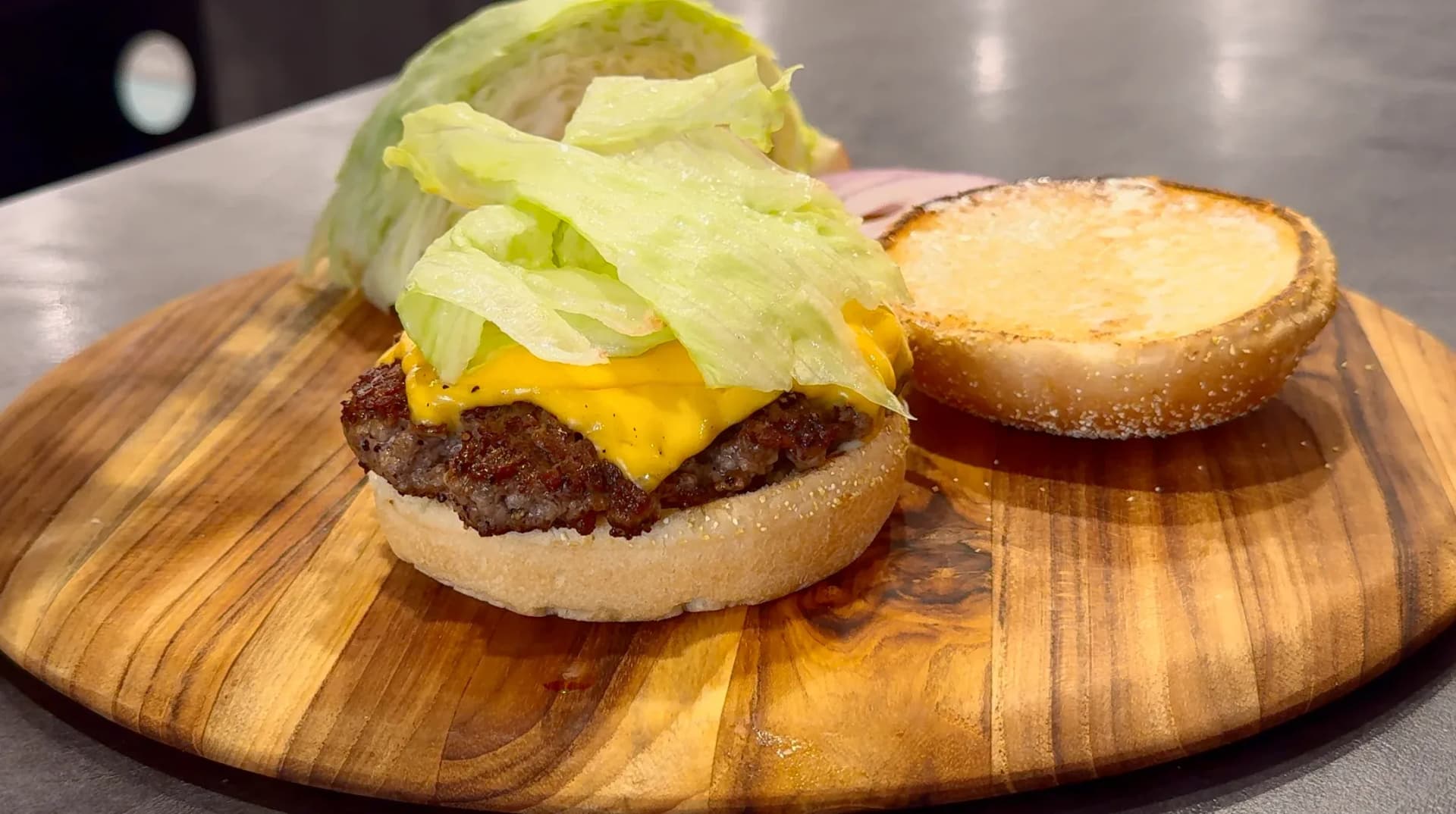 Classic cheeseburger with melted yellow cheese and crisp lettuce on toasted bun, served with toasted bun half on wooden board.