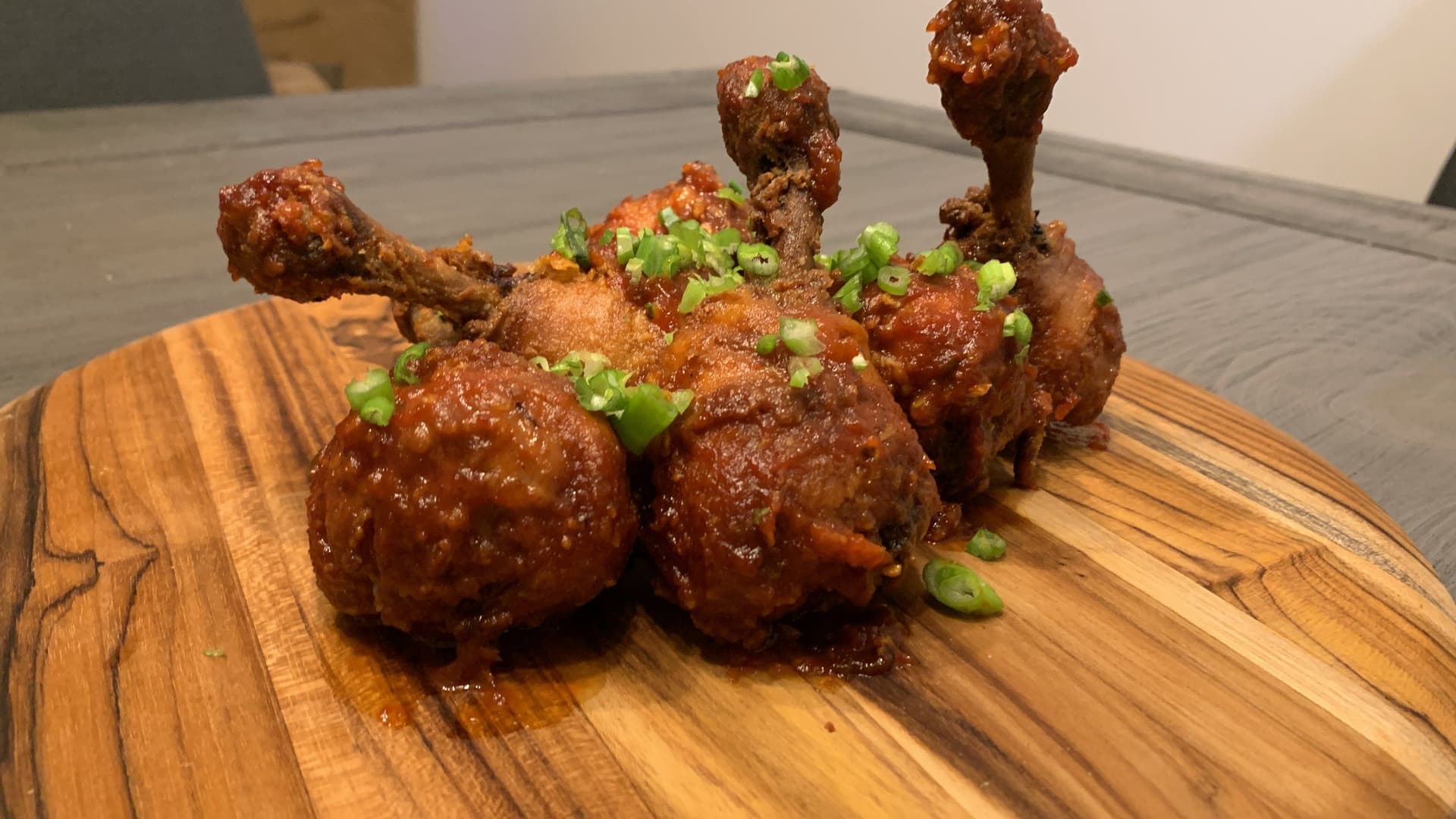 Glazed chicken lollipop drumsticks coated in brown sauce and topped with sliced green onions on a wooden board.