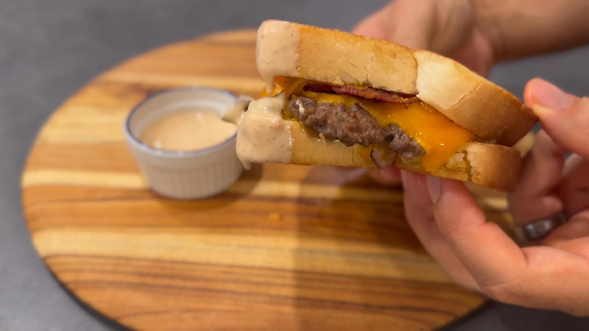 Hand holding a smash burger patty melt with melted cheese on toasted bread, with sauce in background.