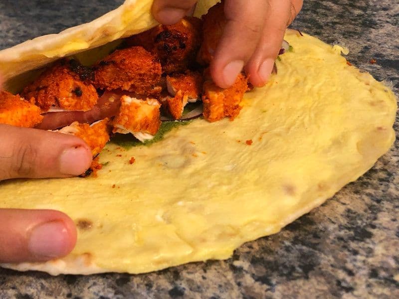 Hands wrapping a yellow paratha around spiced chicken tikka pieces on a granite countertop.