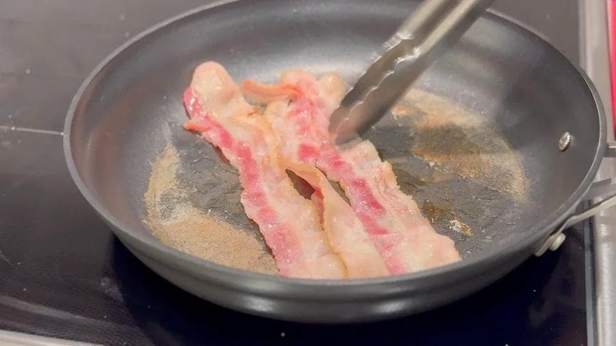 Bacon strips cooking in a dark skillet with a spatula turning them until golden brown.