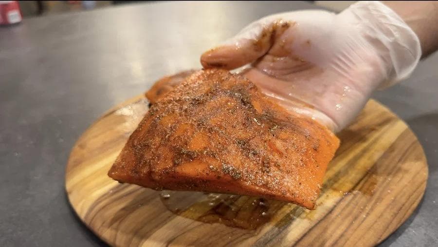 Blackened salmon fillet seasoned with spices on a wooden cutting board, held by a gloved hand.