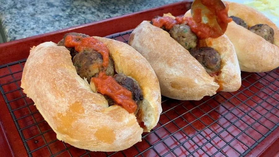 Three Italian meatball subs on a red mesh tray, topped with melted cheese and red marinara sauce on toasted rolls.