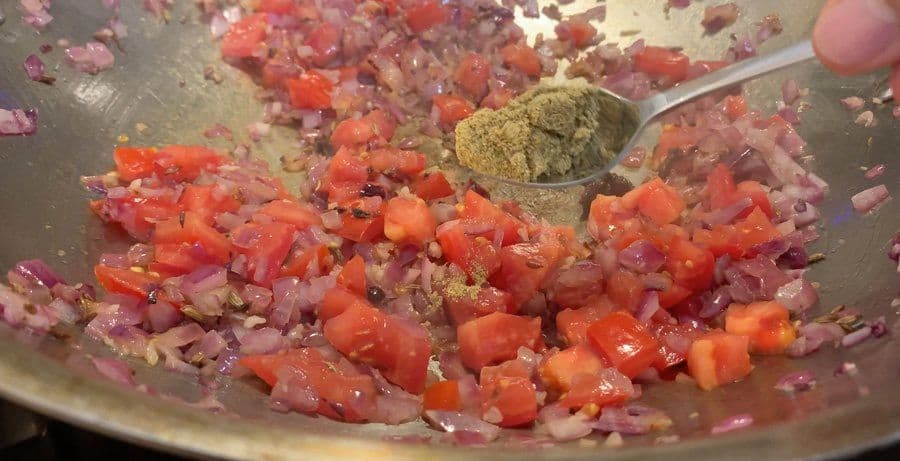 Tomato masala cooking in a pan with diced tomatoes, onions, and spices including ground coriander being added.