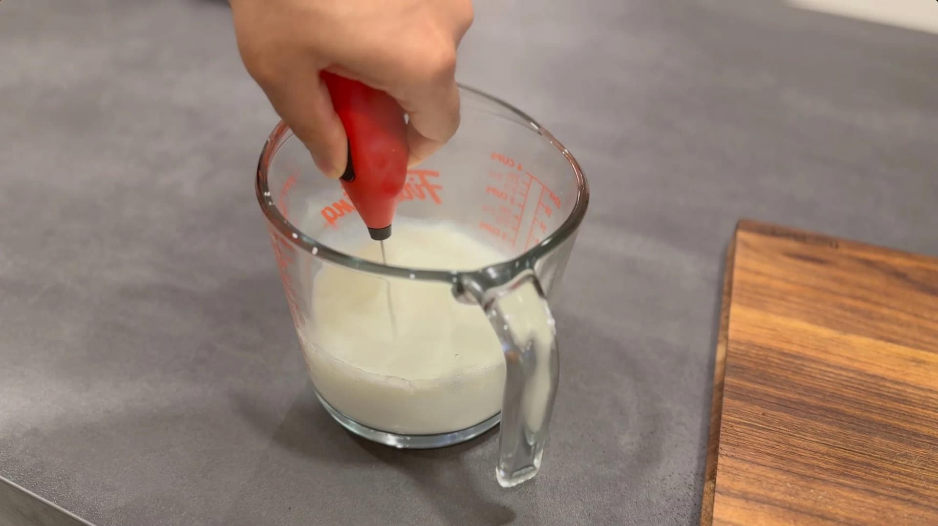 Hand using red electric milk frother in glass measuring cup with white milk.