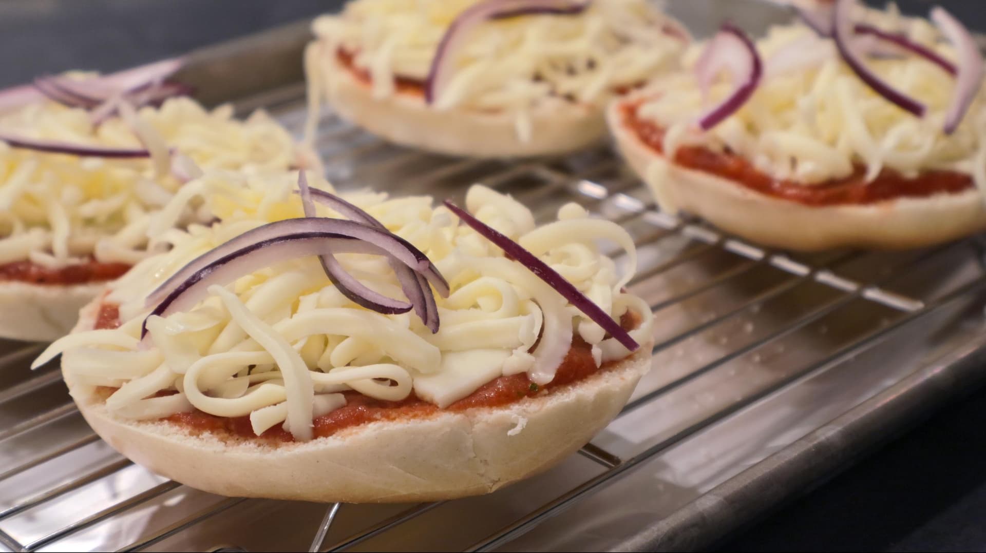 Four assembled bagel pizzas on a wire rack topped with melted mozzarella and red onion slices.
