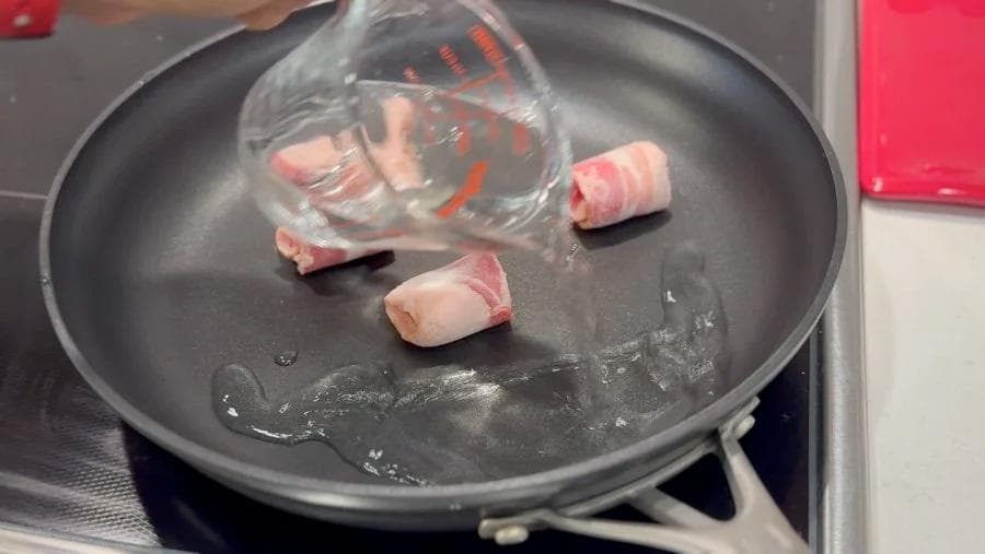 Bacon strips cooking in a dark skillet with water, showing the pan-cooking method for breakfast burrito preparation.