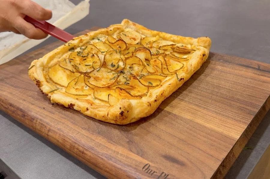 Golden baked puff pastry tart with thinly sliced potatoes and herbs on a wooden board.