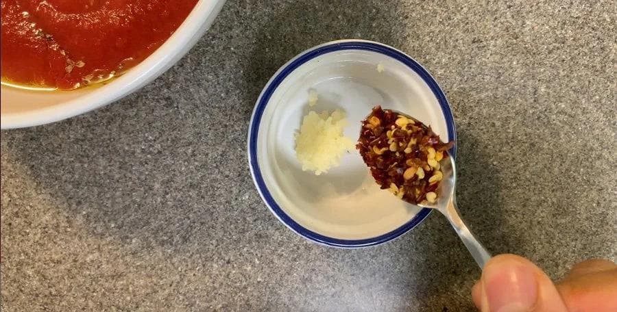 Chilli oil ingredients being mixed in a white bowl with blue rim, including minced garlic and red chili flakes on a spoon.