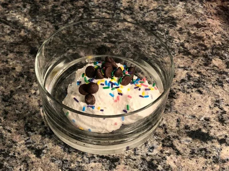 Edible protein chocolate chip cookie dough in a glass bowl topped with chocolate chips and colorful sprinkles on a granite countertop.