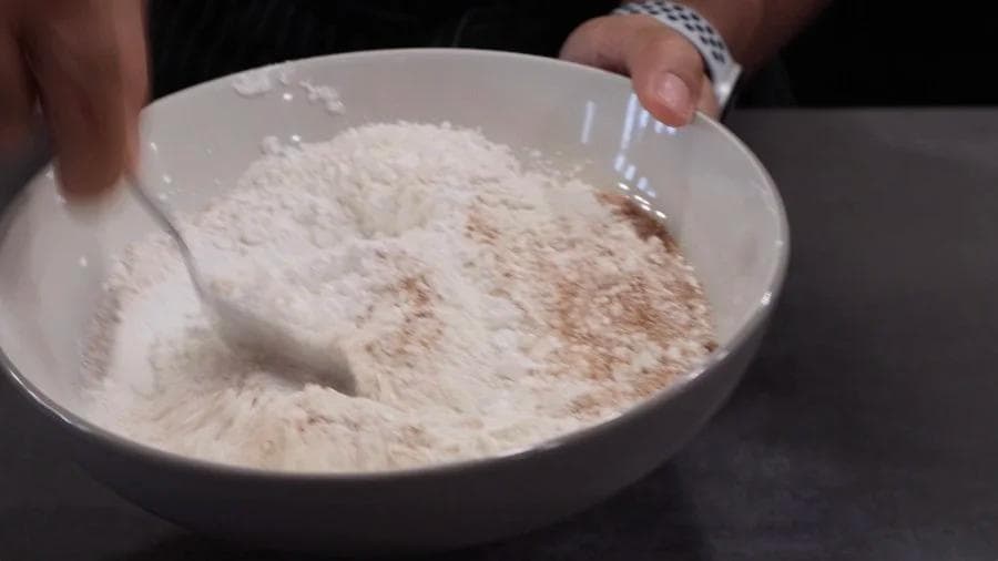 White bowl containing dry breading mix with cinnamon spice sprinkled on top, with hand holding spoon.