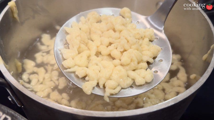 Cooked spätzle being lifted from boiling water with a slotted spoon into a white colander.