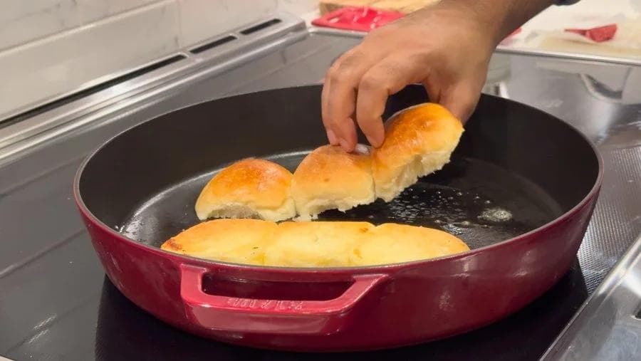 Hand placing toasted dinner roll sliders onto a red pan with melted butter, showing the cooking process.
