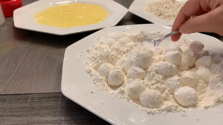 Mozzarella balls being dredged in flour on a white plate, with egg wash and breadcrumbs in separate bowls.