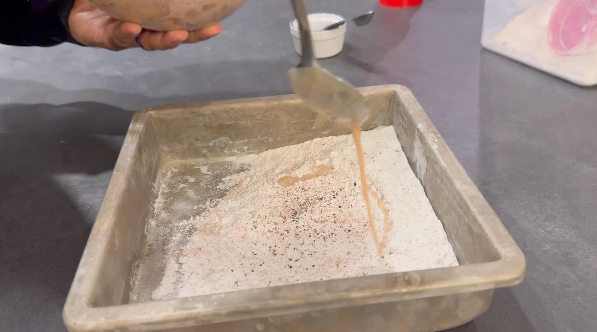 Buttermilk being drizzled into a pan of seasoned flour mixture for fried chicken breading.