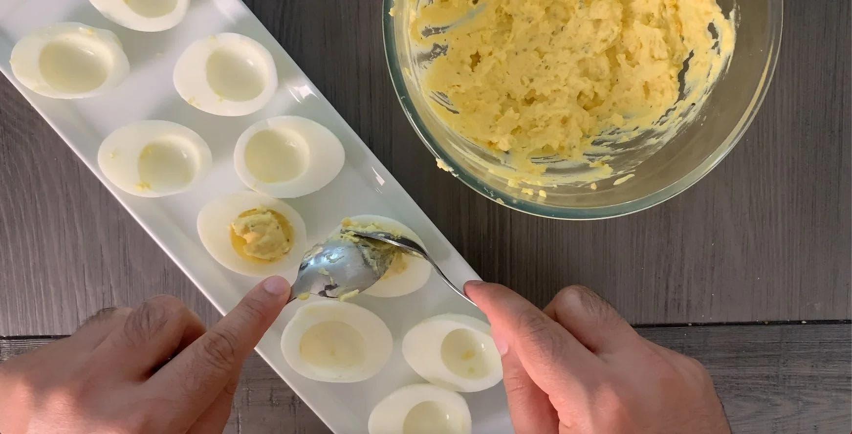 Scooping deviled egg filling into egg white halves using two spoons over a muffin tin.