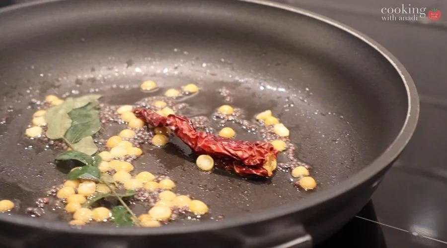 Tempering spices including dried red chili, chana dal, mustard seeds, and curry leaves sizzling in a dark pan.