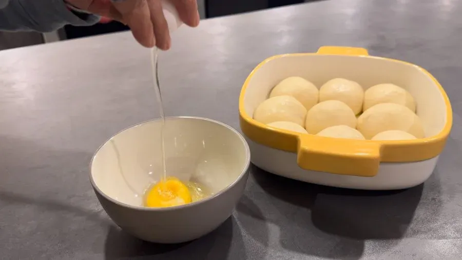 Preparing egg wash by whisking egg and water in a white bowl next to a yellow egg holder with raw eggs.