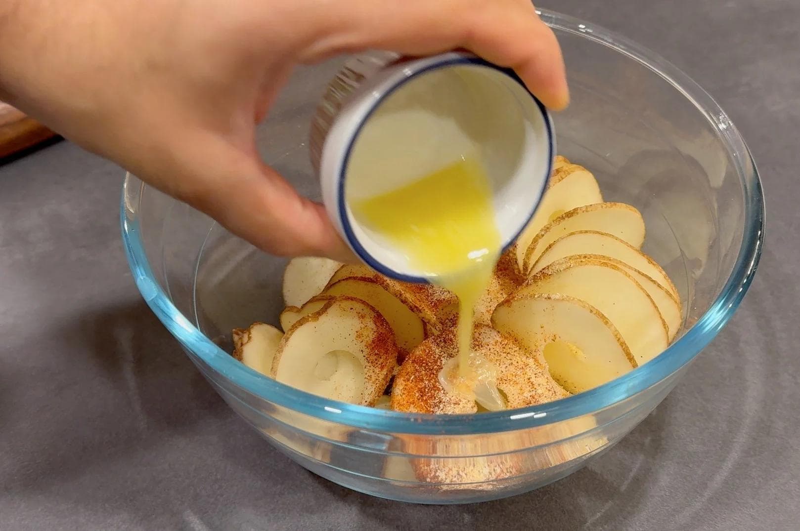 Hand drizzling melted butter over thinly sliced potatoes seasoned with spices in a glass bowl