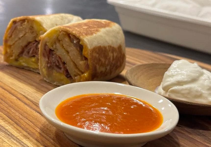 Sliced breakfast burrito with chorizo and egg filling, served with salsa and sour cream on wooden board.
