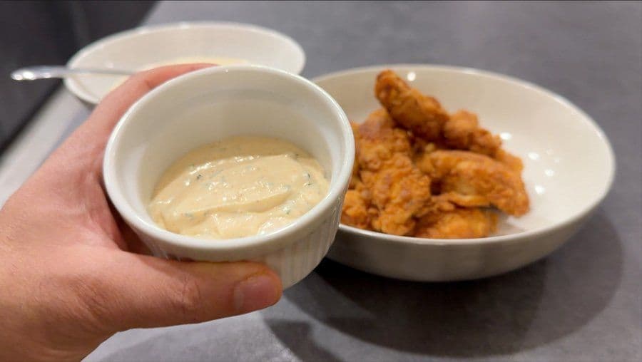 Hand holding a small bowl of creamy ranch dip above a plate of fried chicken pieces.
